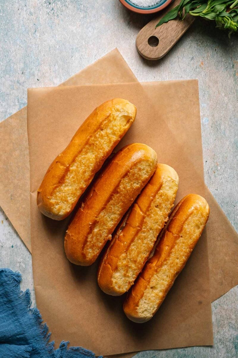 Four golden-brown split top hot dog buns are arranged in a row on parchment paper, set on a light textured surface with a blue cloth and herbs partially visible.