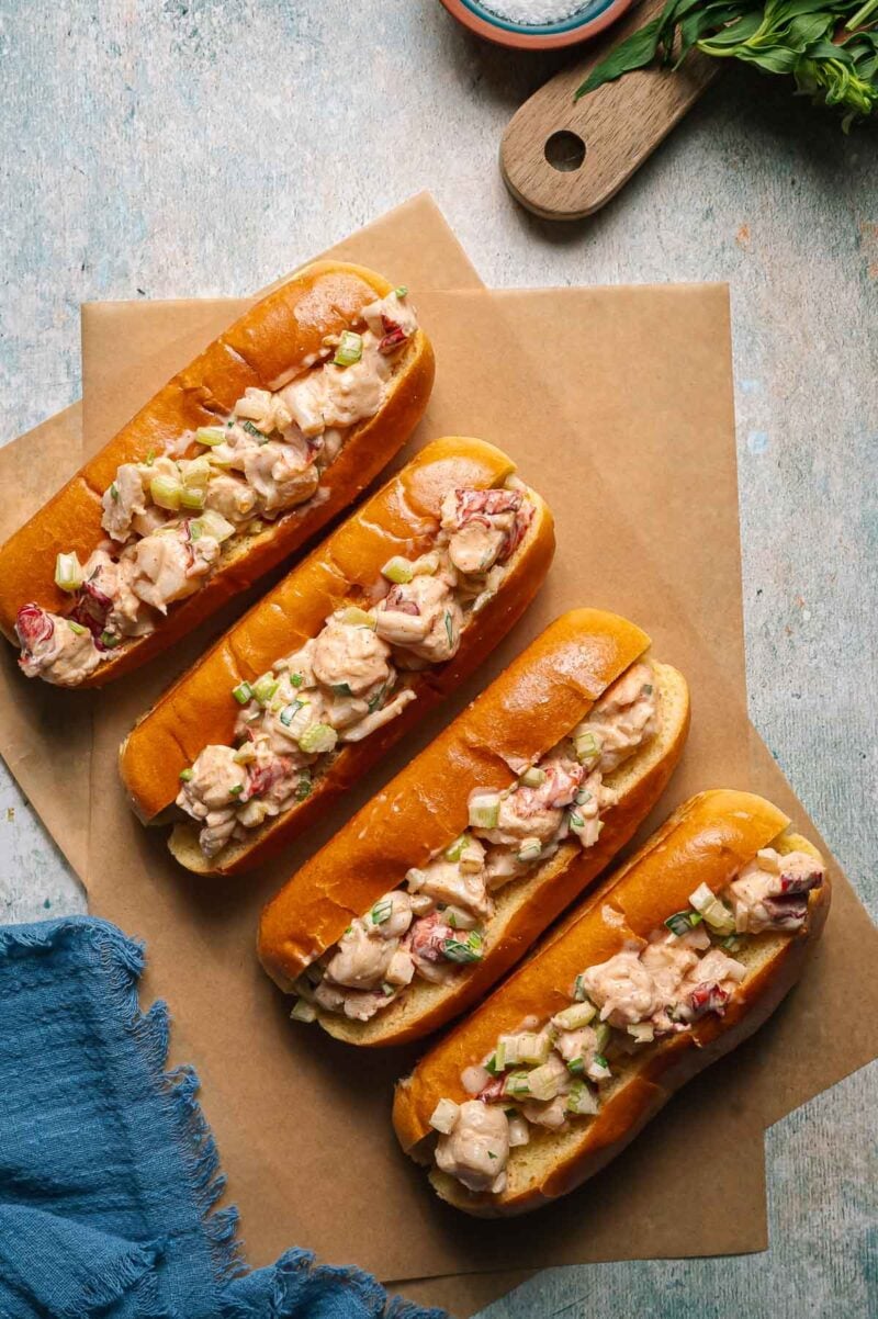 Four maine lobster rolls filled with lobster salad are arranged on parchment paper atop a light-colored surface.