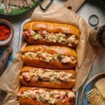 Four split top toasted buns filled with creamy lobster salad, garnished with chopped vegetables, are arranged on parchment paper next to chips and seasonings on a rustic table.