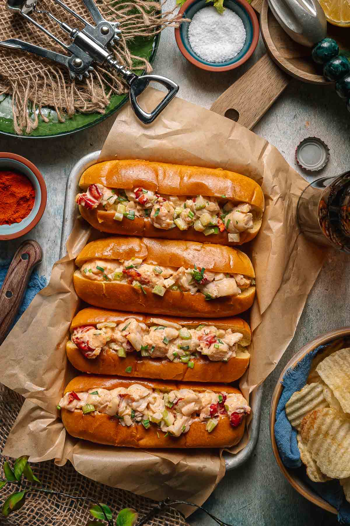 Four split top toasted buns filled with creamy lobster salad, garnished with chopped vegetables, are arranged on parchment paper next to chips and seasonings on a rustic table.