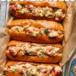 Overhead view of four Maine lobster rolls on a metal tray, filled with lobster meat and diced vegetables, set on a wooden surface. Text overlay with recipe website information.