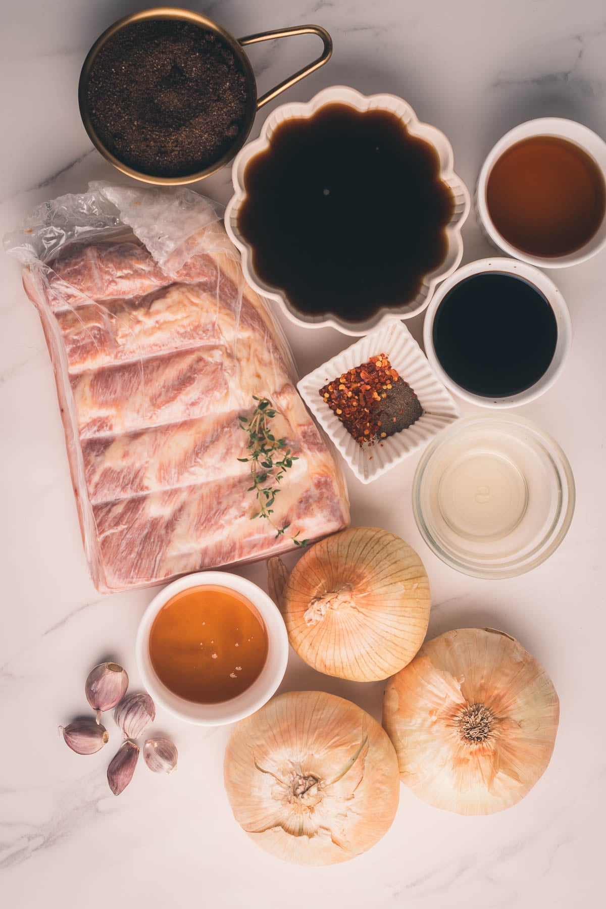 A slab of raw pork belly, onions, garlic cloves, and small bowls of brown sugar, vinegar, coffee, honey, spices, and liquids arranged on a white marble surface.
