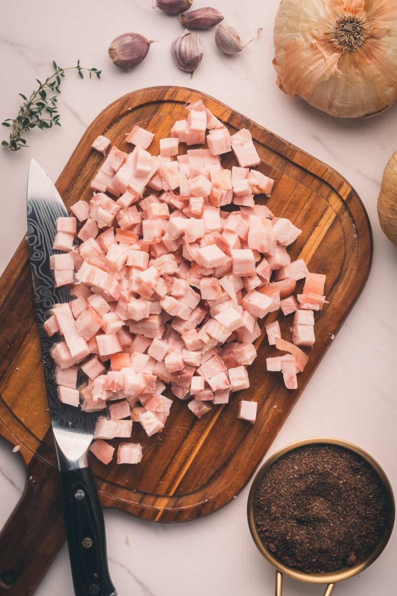 A wooden cutting board with diced bacon, a knife, onions, garlic cloves, thyme, and a measuring cup of brown sugar on a white surface.