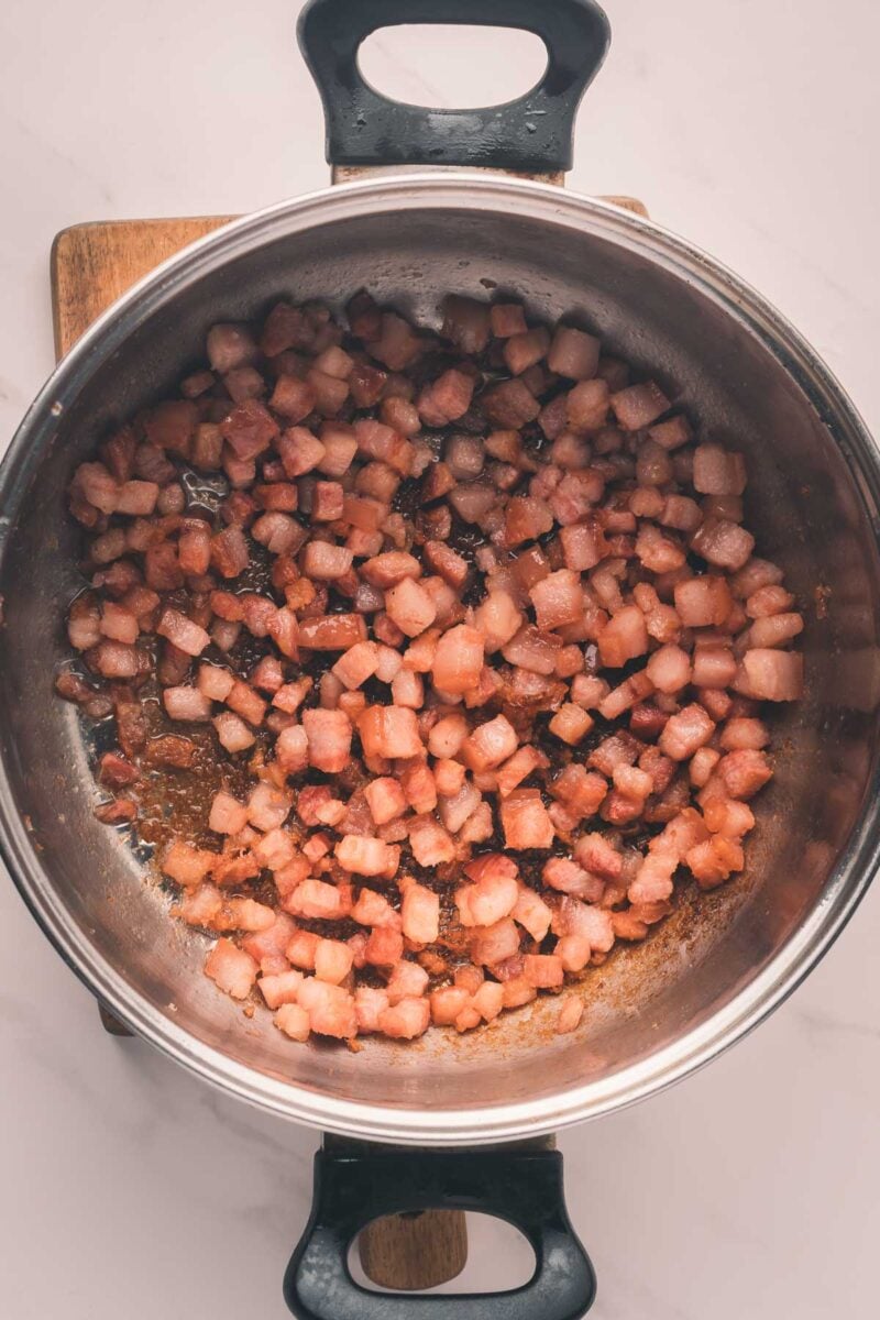 Diced bacon pieces are cooking and browning in a stainless steel pot on a wooden trivet, viewed from above.