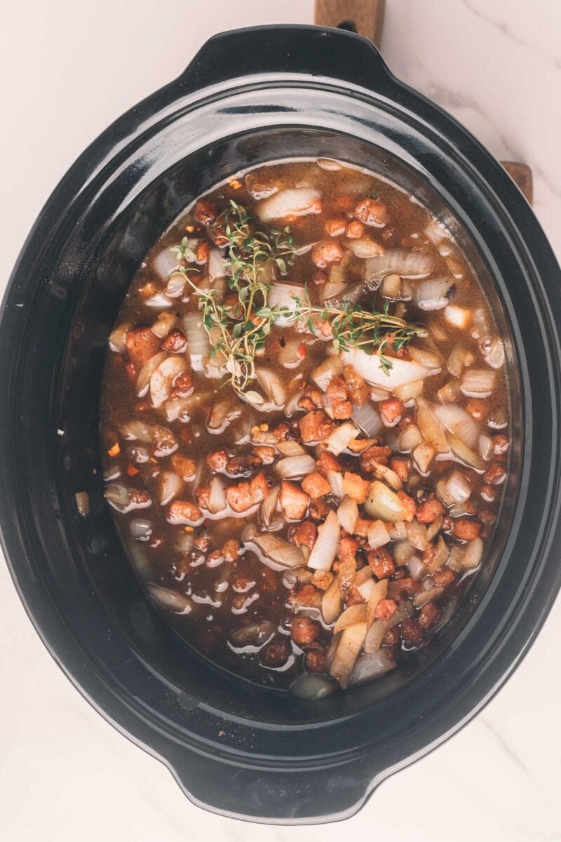 A slow cooker filled with a mixture of chopped onions, diced bacon, coffee, and herbs, ready to cook.