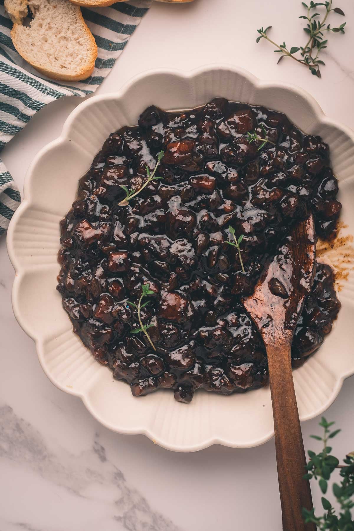 A white scalloped bowl filled with dark, chunky bacon jam, garnished with herbs, with a wooden spoon resting inside and bread slices nearby.