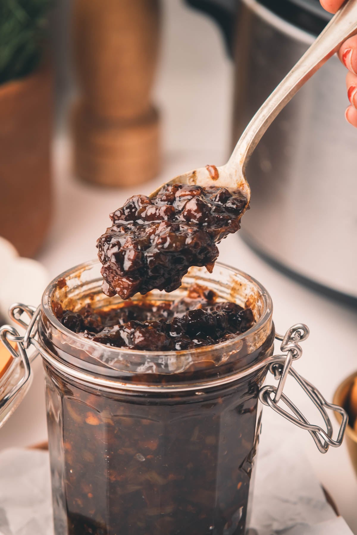 A spoonful of dark, chunky bacon jam being held over an open glass jar filled with the same chutney.
