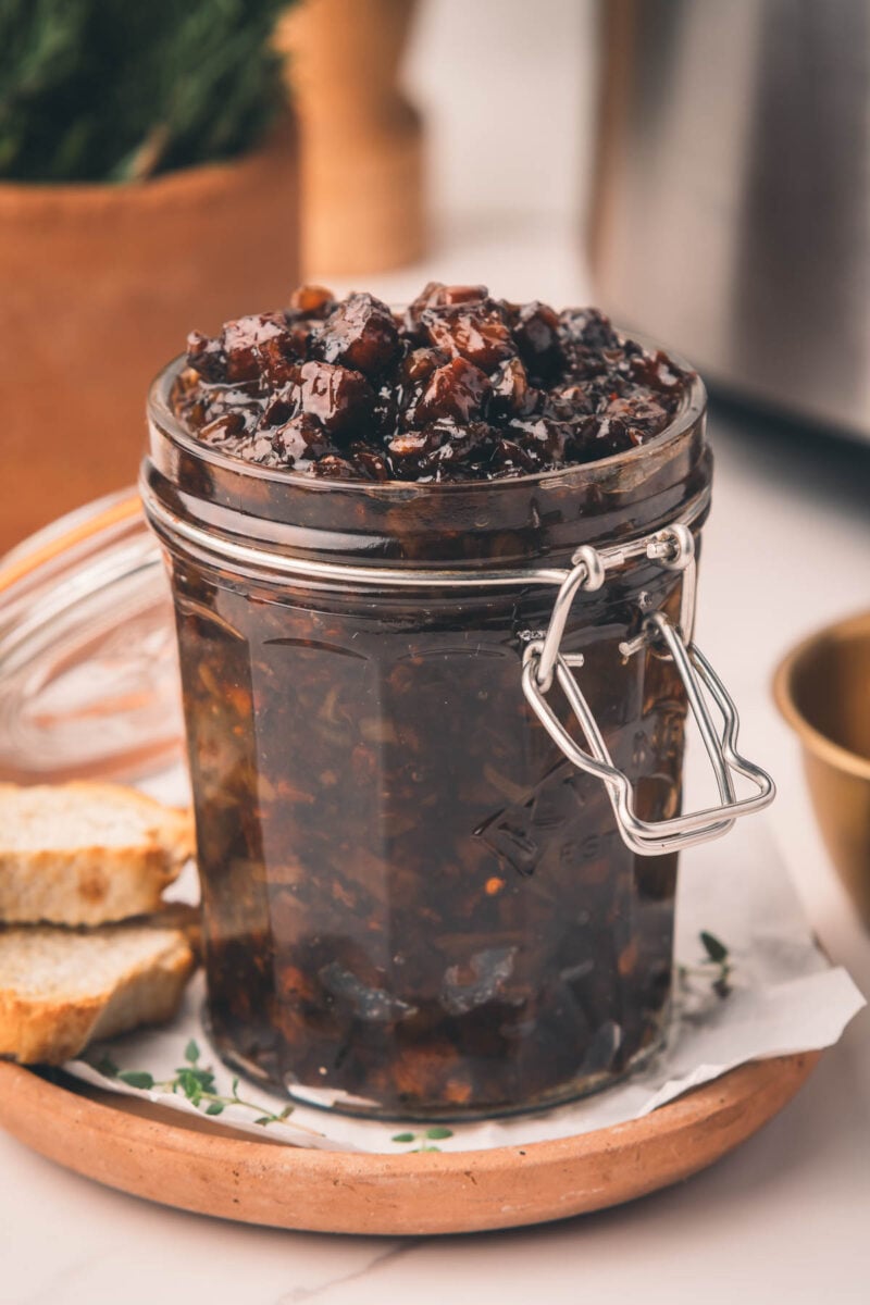 A glass jar filled with bacon jam sits on a plate with two slices of bread beside it.
