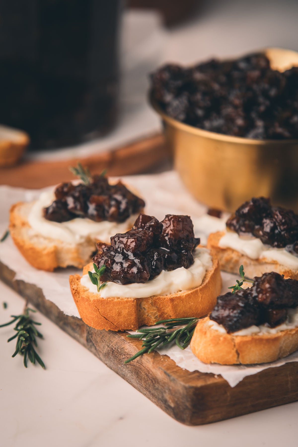 Three pieces of toasted bread topped with a white spread and a chunky onion bacon jam are arranged on a wooden board, garnished with fresh herbs. A bowl of the topping is in the background.