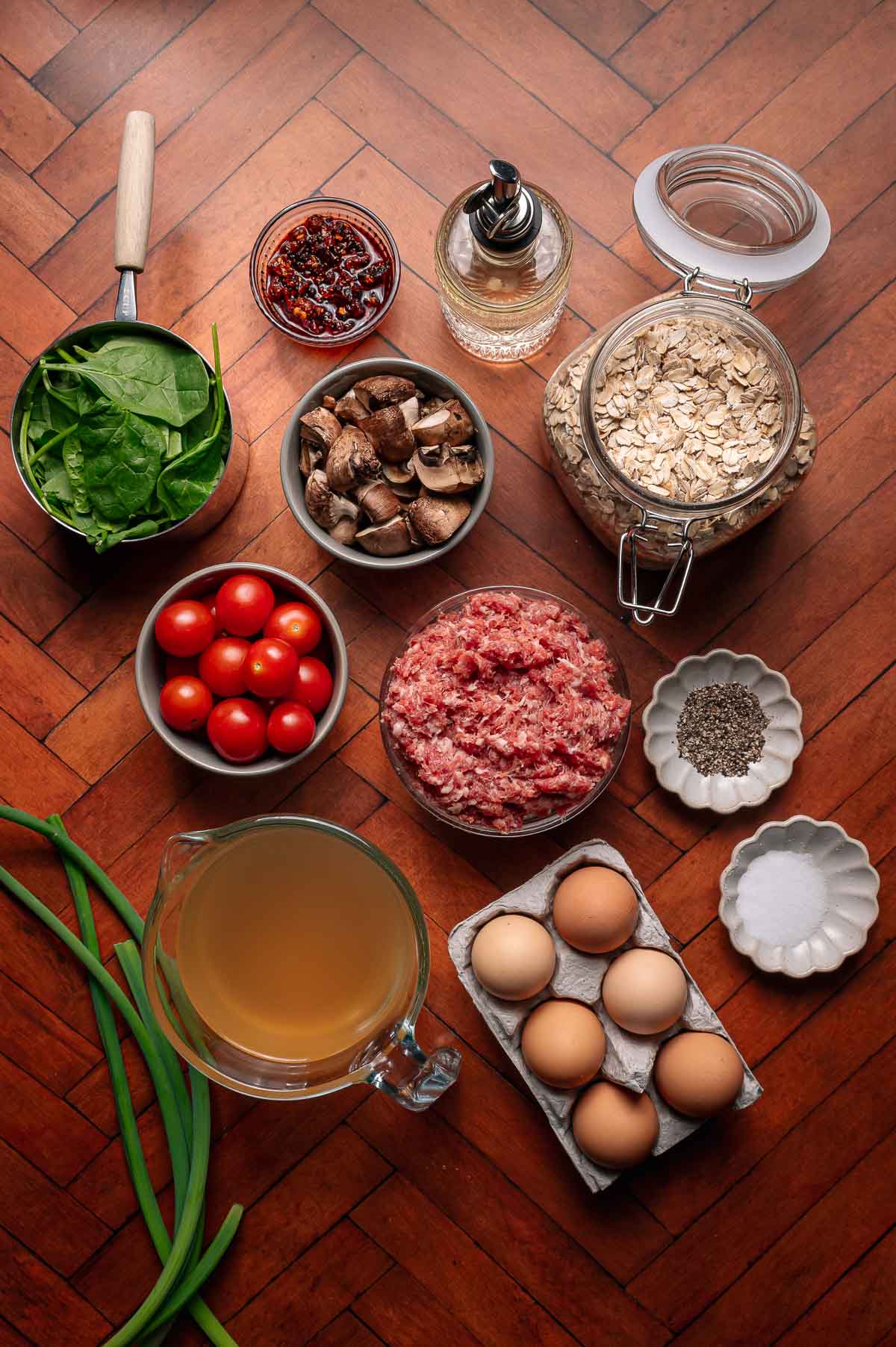 A top-down view of raw ingredients on a wooden surface, including spinach, cherry tomatoes, mushrooms, oats, ground meat, eggs, broth, green onions, seasonings, and oil.