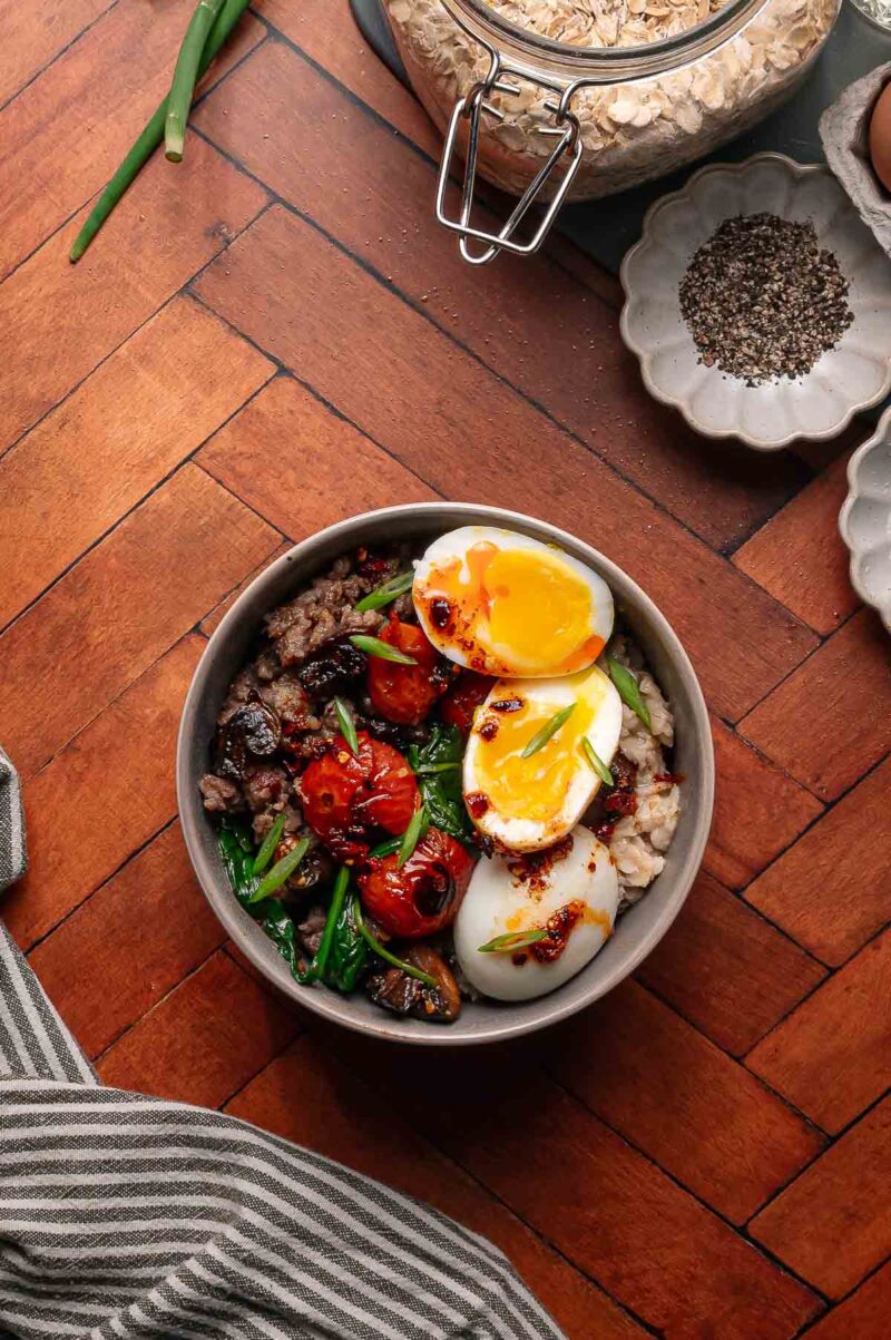 A bowl of oatmeal topped with sautéed vegetables, ground meat, tomatoes, green onions, and two soft-boiled eggs with runny yolks, placed on a wooden surface.