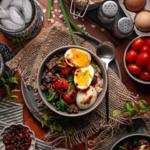 A bowl of rice topped with soft-boiled eggs, sautéed vegetables, cherry tomatoes, and chili oil, surrounded by fresh ingredients and kitchen utensils on a rustic table.