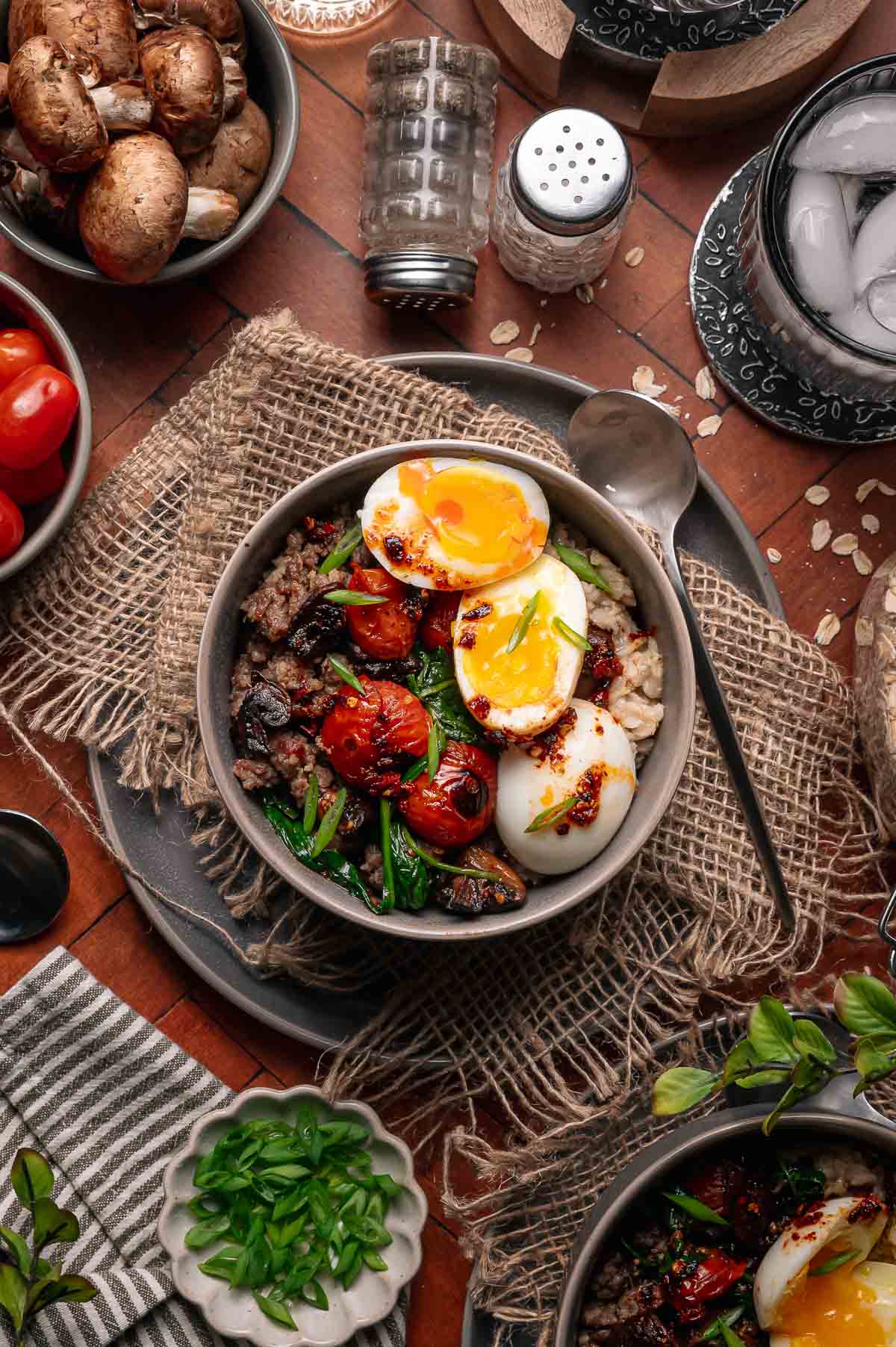 A bowl of rice topped with halved soft-boiled eggs, roasted cherry tomatoes, greens, ground meat, and chili oil, surrounded by mushrooms, tomatoes, and seasonings on a rustic table.