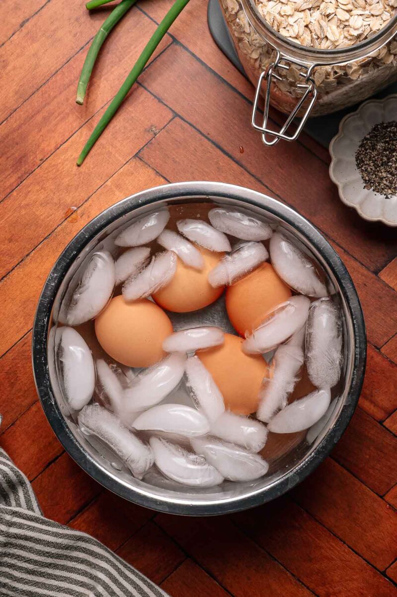 Three brown eggs are submerged in a metal bowl filled with ice water and ice cubes, placed on a wooden floor next to oats, green onions, and ground black pepper.