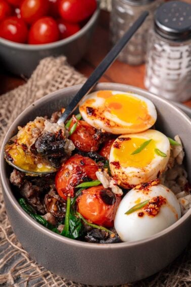 A bowl of roasted tomatoes, sautéed greens, ground meat, and halved soft-boiled eggs, garnished with green onions. A spoon rests inside the bowl.