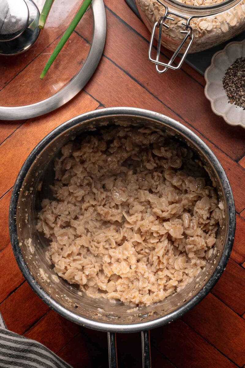 A stainless steel pot filled with cooked oatmeal sits on a wooden surface, surrounded by a jar of oats, a plate of black pepper, green onions, and a pot lid.