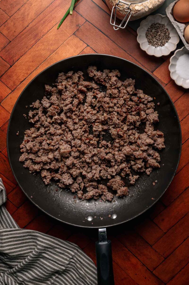 Cooked ground beef in a black skillet sits on a wooden surface, surrounded by eggs, pepper, and a striped cloth.