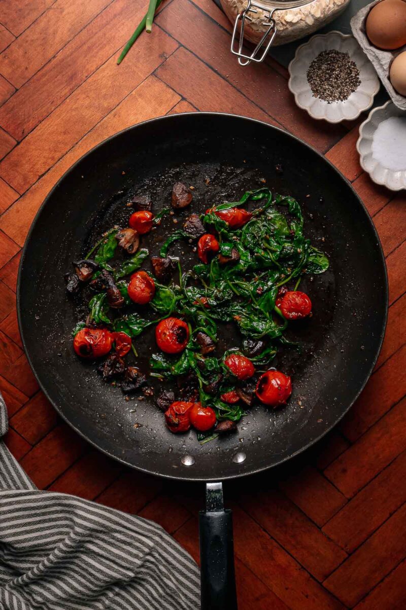 A black skillet on a wooden floor contains sautéed cherry tomatoes, spinach, and mushrooms. Eggs, black pepper, and a dish towel are nearby.
