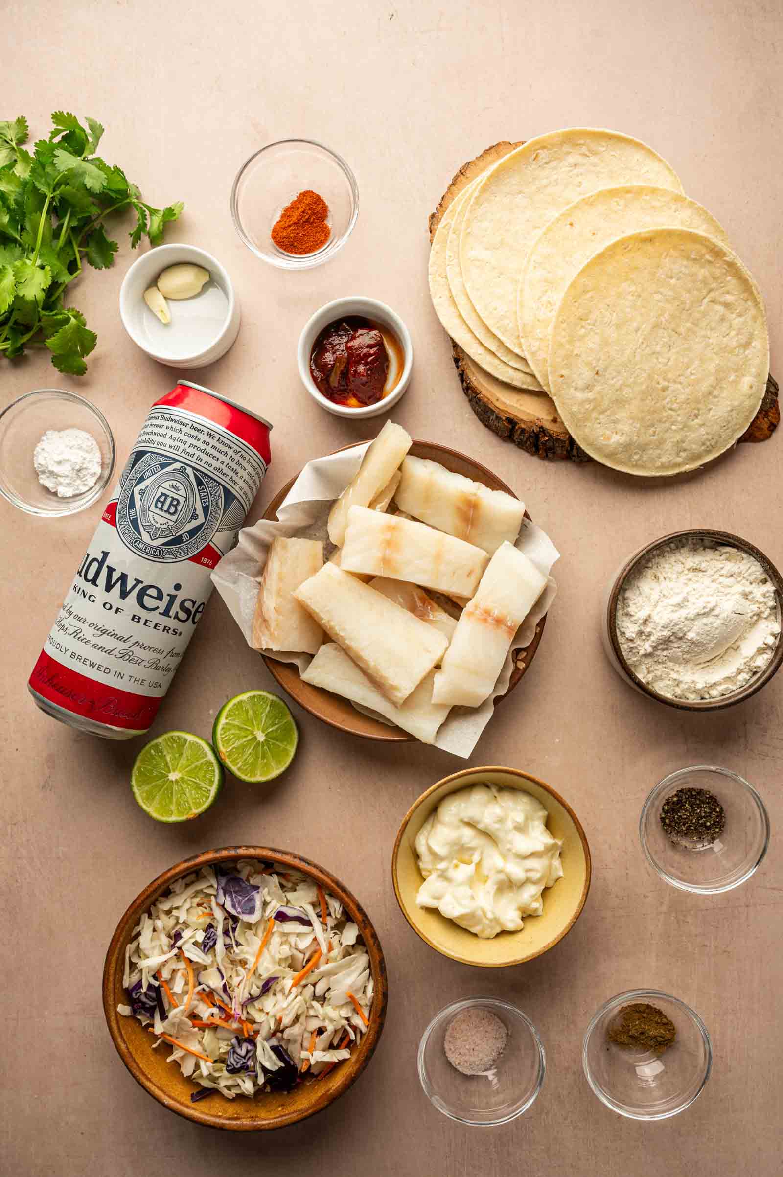 A variety of ingredients for fish tacos are arranged on a table, including tortillas, raw fish, a can of beer, lime, coleslaw, seasonings, flour, mayonnaise, and fresh cilantro.