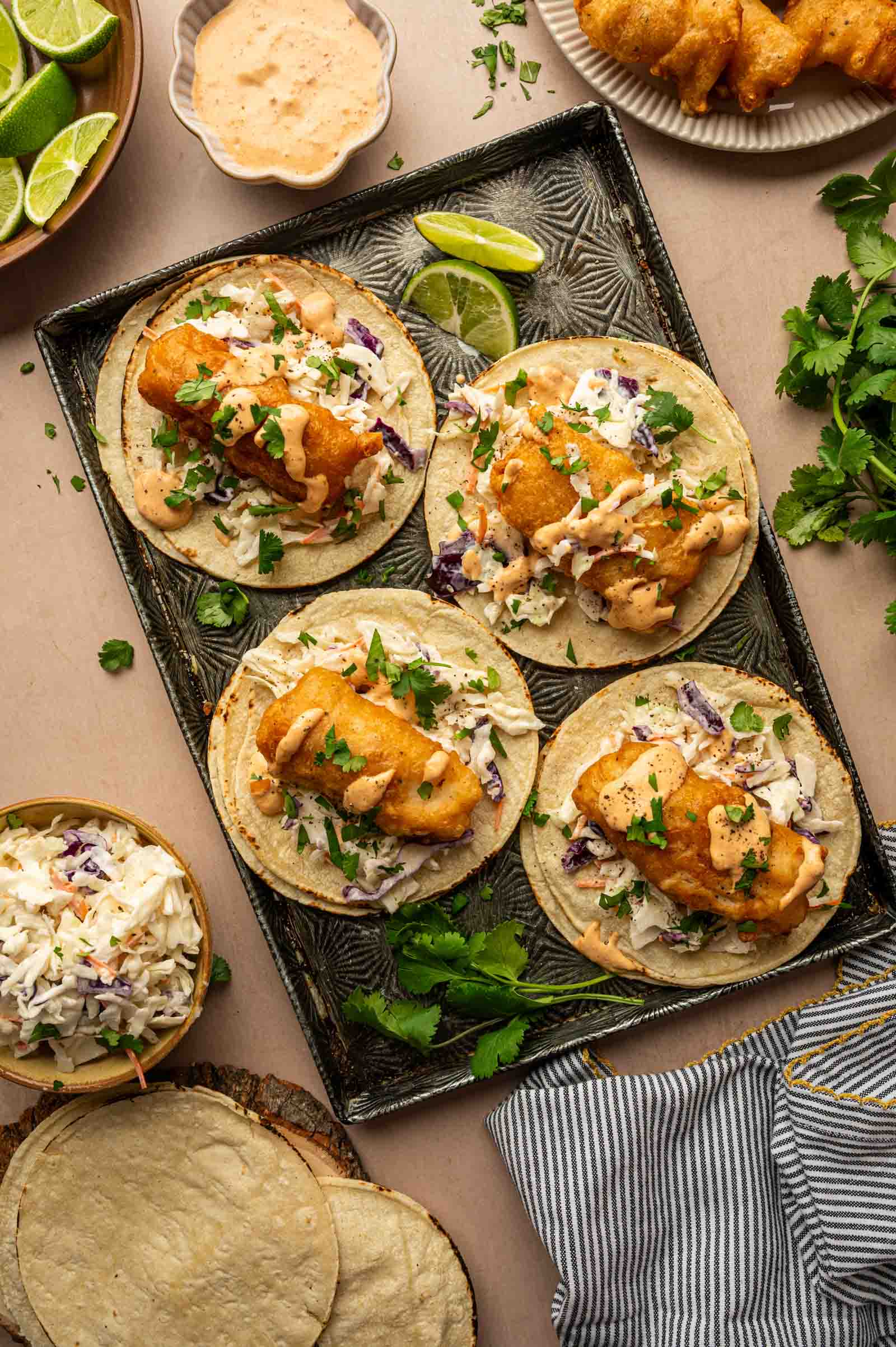 Four fish tacos with fried fish, slaw, sauce, and cilantro are arranged on a tray, with lime wedges and extra toppings on the side.