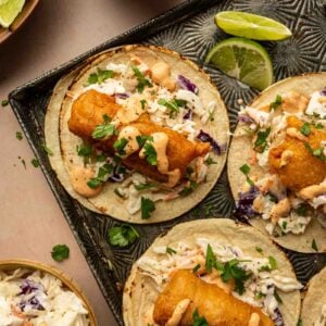 Three baja fish tacos topped with slaw, cilantro, and creamy sauce are arranged on a tray with lime wedges. A bowl of slaw and a bowl of sauce are nearby.