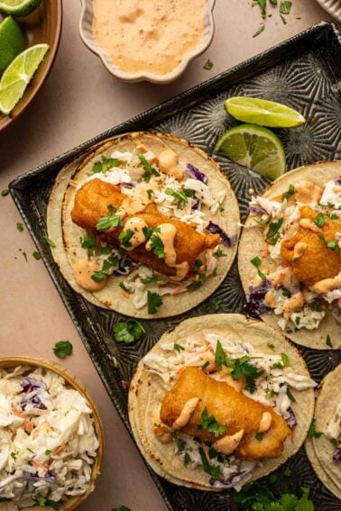 Three baja fish tacos topped with slaw, cilantro, and creamy sauce are arranged on a tray with lime wedges. A bowl of slaw and a bowl of sauce are nearby.
