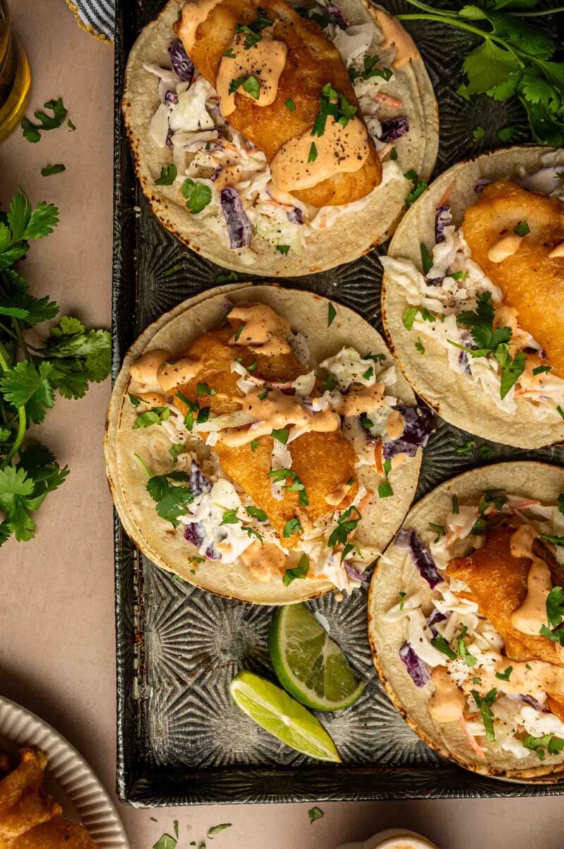 Tray of fish tacos topped with slaw, chopped herbs, and creamy sauce, served with lime wedges and fresh cilantro on the side.