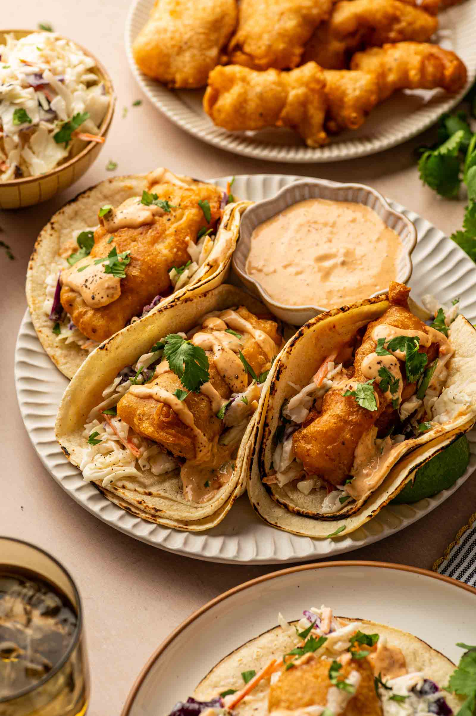 Three baja fish tacos topped with slaw and chiptole sauce sit on a plate next to a bowl of dipping sauce, with fried fish and coleslaw in the background.