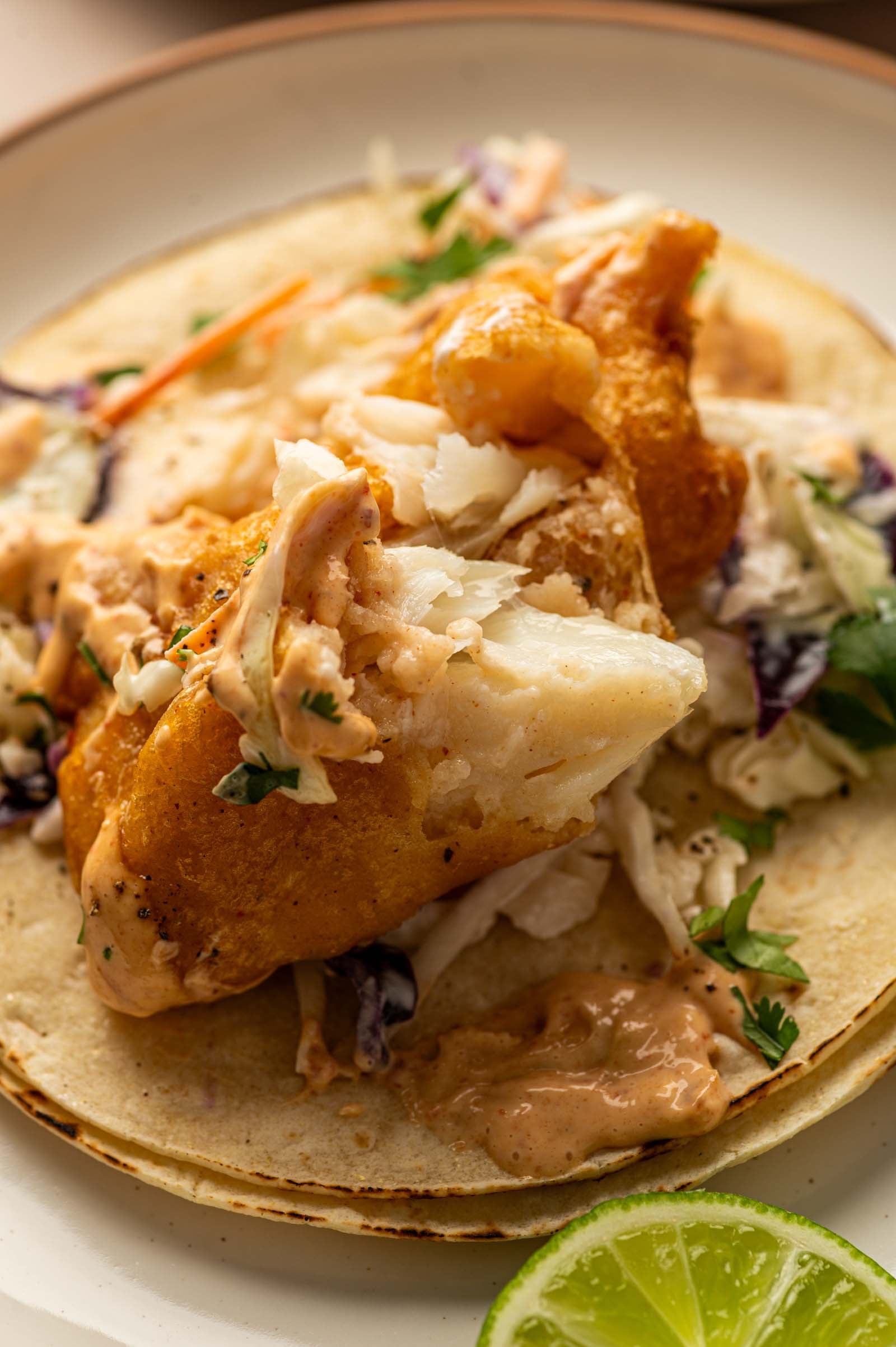 A fish taco with battered fish, shredded cabbage, sauce, and herbs on a corn tortilla, served with a slice of lime on the side.