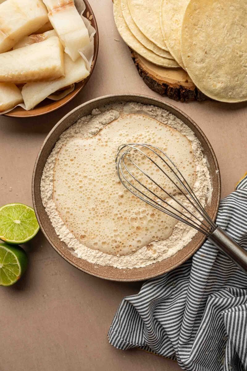 A bowl of bubbling batter with a whisk rests on flour, surrounded by tortillas, lime halves, sliced waxy ingredient, and a striped cloth.