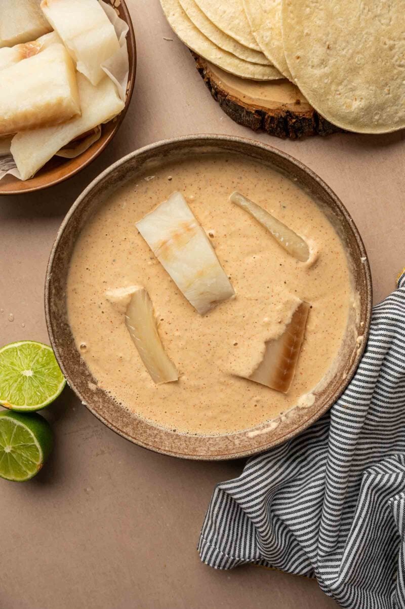 A brown bowl filled with creamy batter and pieces of white fish, surrounded by tortillas, a halved lime, and a striped cloth.