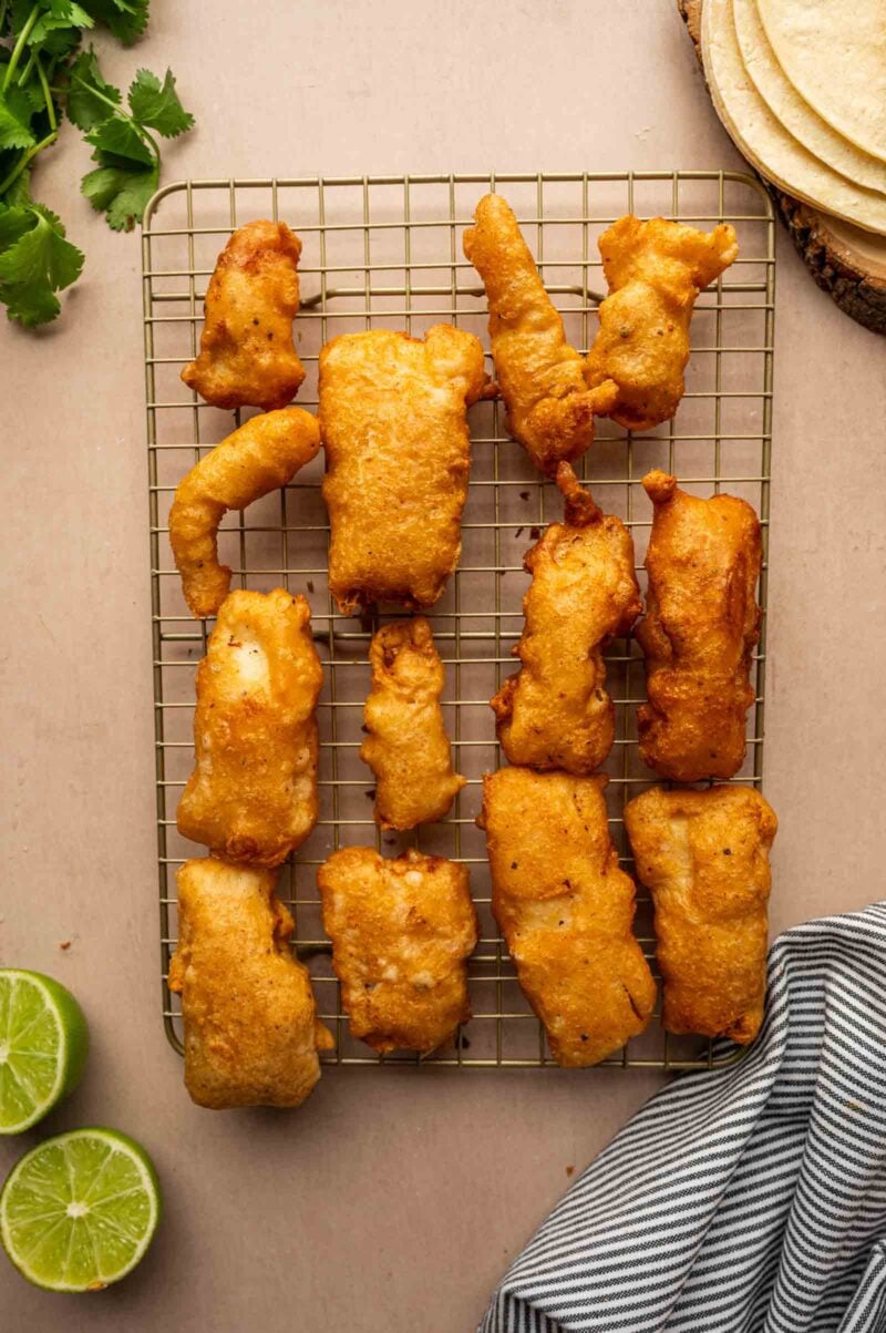Golden battered fish pieces arranged on a cooling rack, with lime halves, cilantro, tortillas, and a striped cloth nearby.