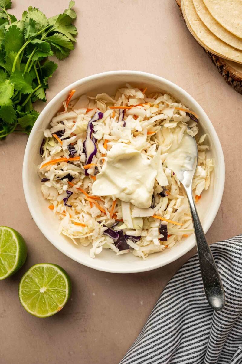 A bowl of coleslaw mix with a dollop of mayonnaise and a spoon, surrounded by lime halves, cilantro, tortillas, and a striped cloth.