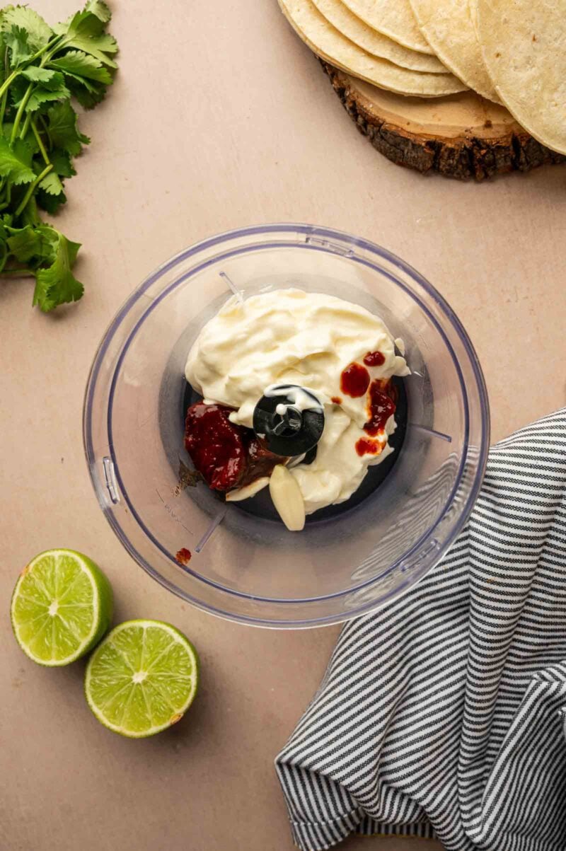 A food processor bowl with mayonnaise, chipotle peppers, garlic, and sauce; nearby are cilantro, lime halves, tortillas, and a striped cloth.