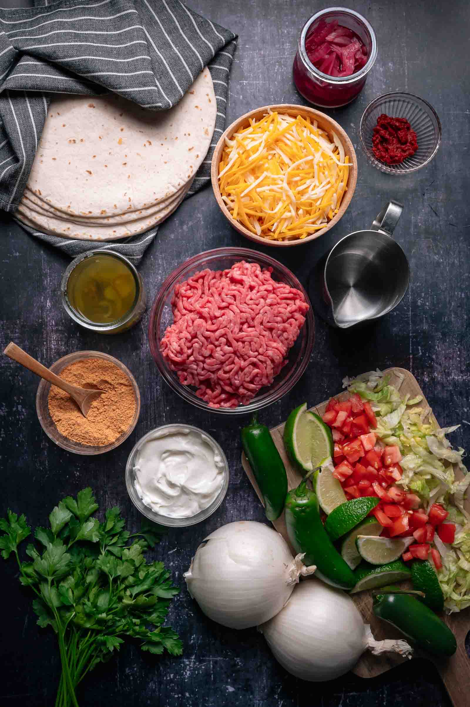 Overhead view of taco ingredients including ground beef, shredded cheese, tortillas, onion, lettuce, tomatoes, jalapeños, lime wedges, sour cream, pickled onions, seasonings, and olive oil.