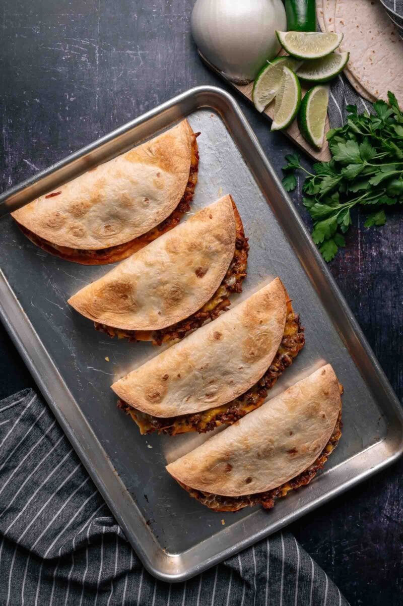 A baking sheet with four folded, baked tacos filled with meat, set on a dark surface next to sliced limes, tortillas, parsley, and an onion.