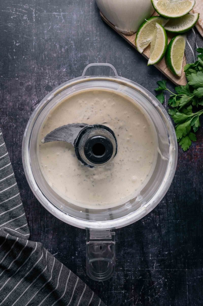 Food processor bowl filled with creamy jalapeno sour cream sauce, placed on a dark countertop near lime wedges, herbs, and a striped kitchen towel.