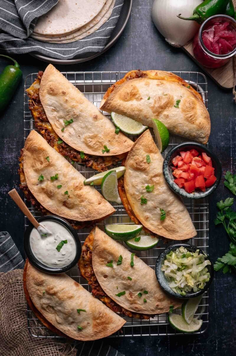 Six baked crispy tacos filled with ground beef are arranged on a cooling rack, served with lime wedges, diced tomatoes, shredded lettuce, and a bowl of white sauce.