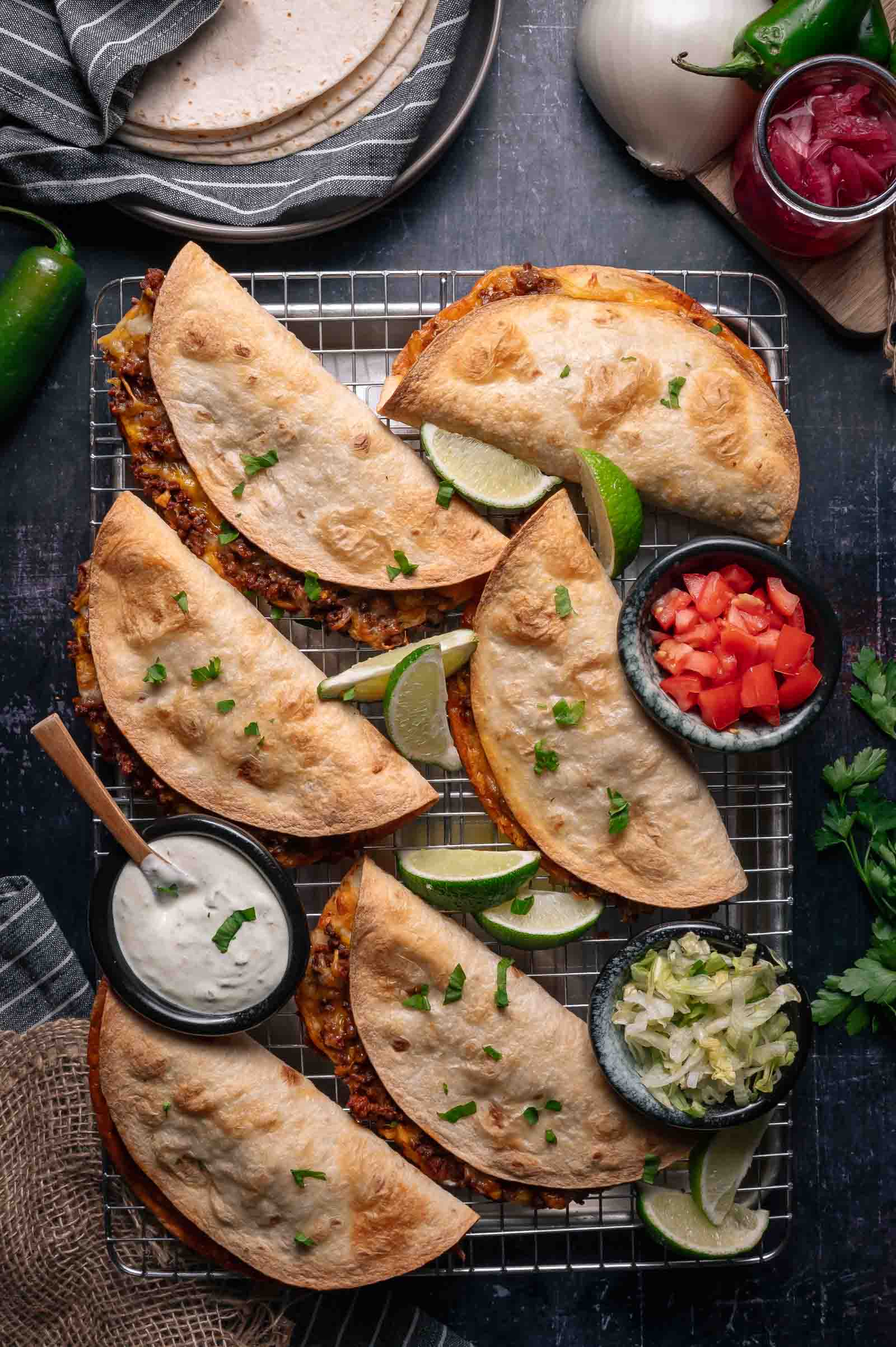 Six baked crispy tacos filled with ground beef are arranged on a cooling rack, served with lime wedges, diced tomatoes, shredded lettuce, and a bowl of white sauce.