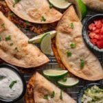 Five baked tacos with ground meat and melted cheese on a cooling rack, garnished with lime wedges, with bowls of sour cream, chopped tomatoes, and shredded lettuce on the side.