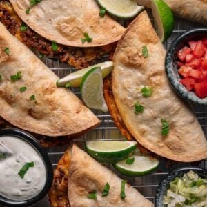 Five baked tacos with ground meat and melted cheese on a cooling rack, garnished with lime wedges, with bowls of sour cream, chopped tomatoes, and shredded lettuce on the side.