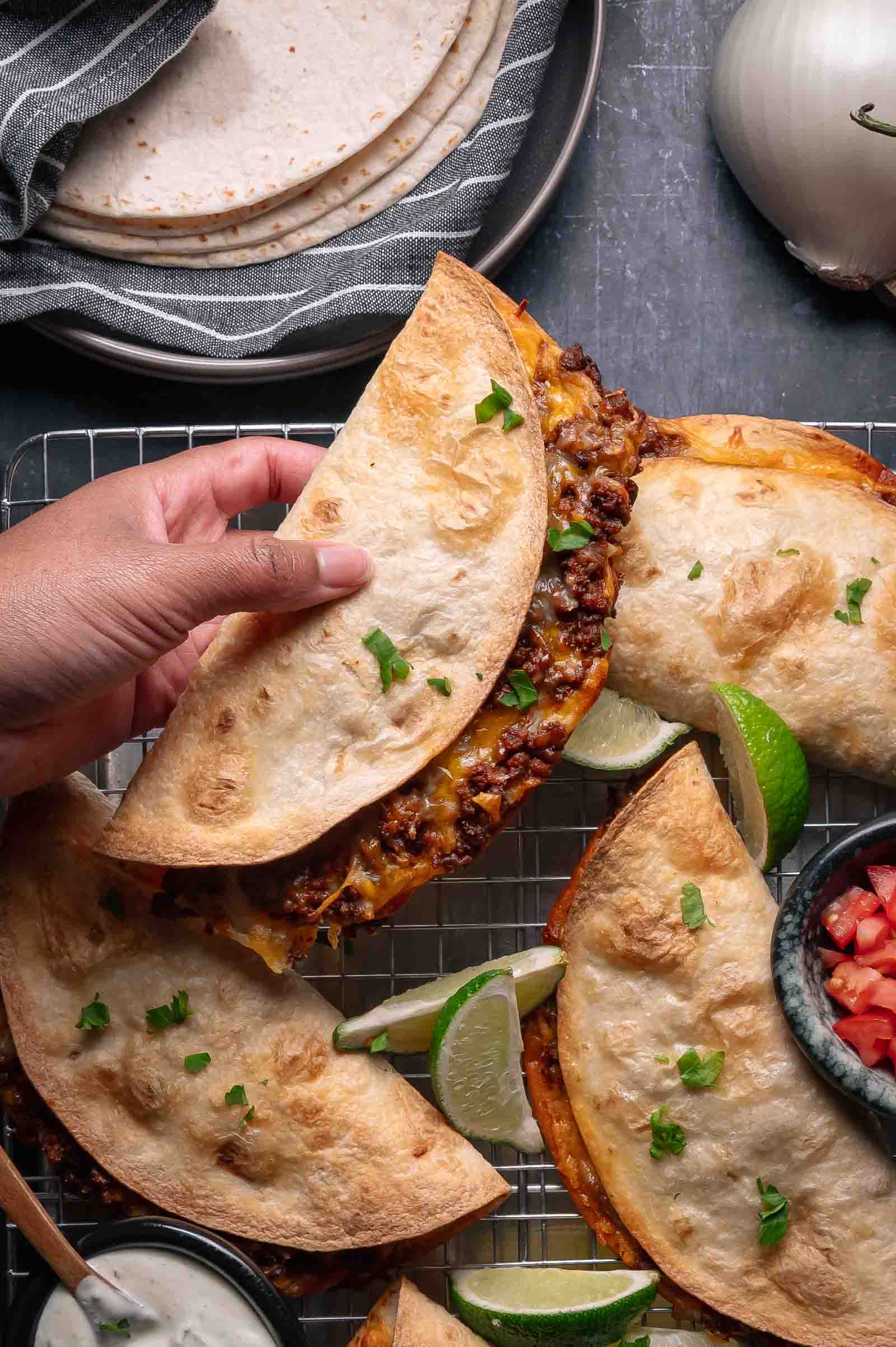 A hand holds a crispy baked beef taco filled with ground beef and garnished with chopped herbs; other quesadillas, lime wedges, and bowls of salsa and sauce are nearby.