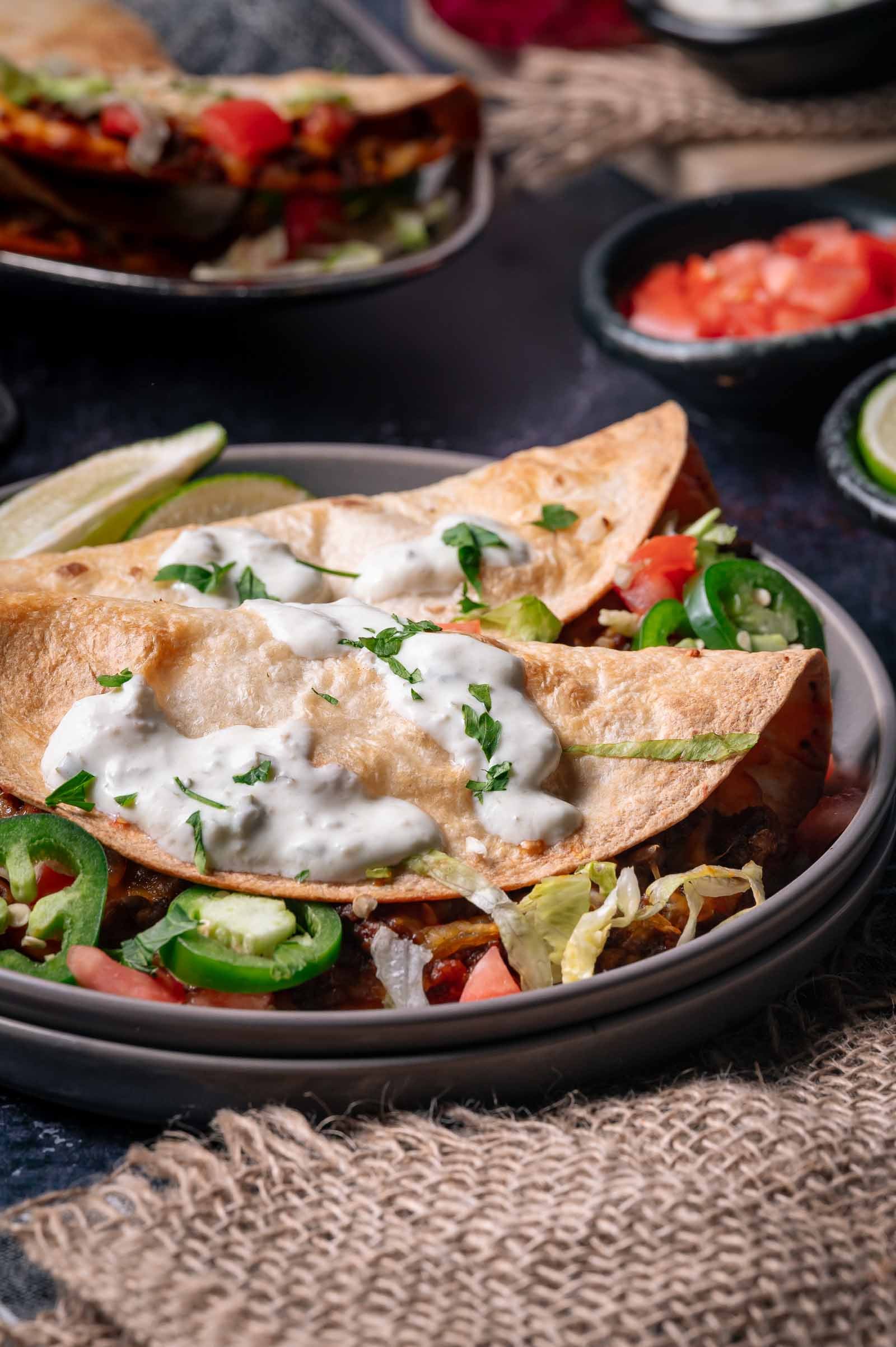 Two crispy tacos filled with lettuce, tomato, and sliced jalapeños, topped with a white creamy sauce, served on a gray plate with lime wedges.