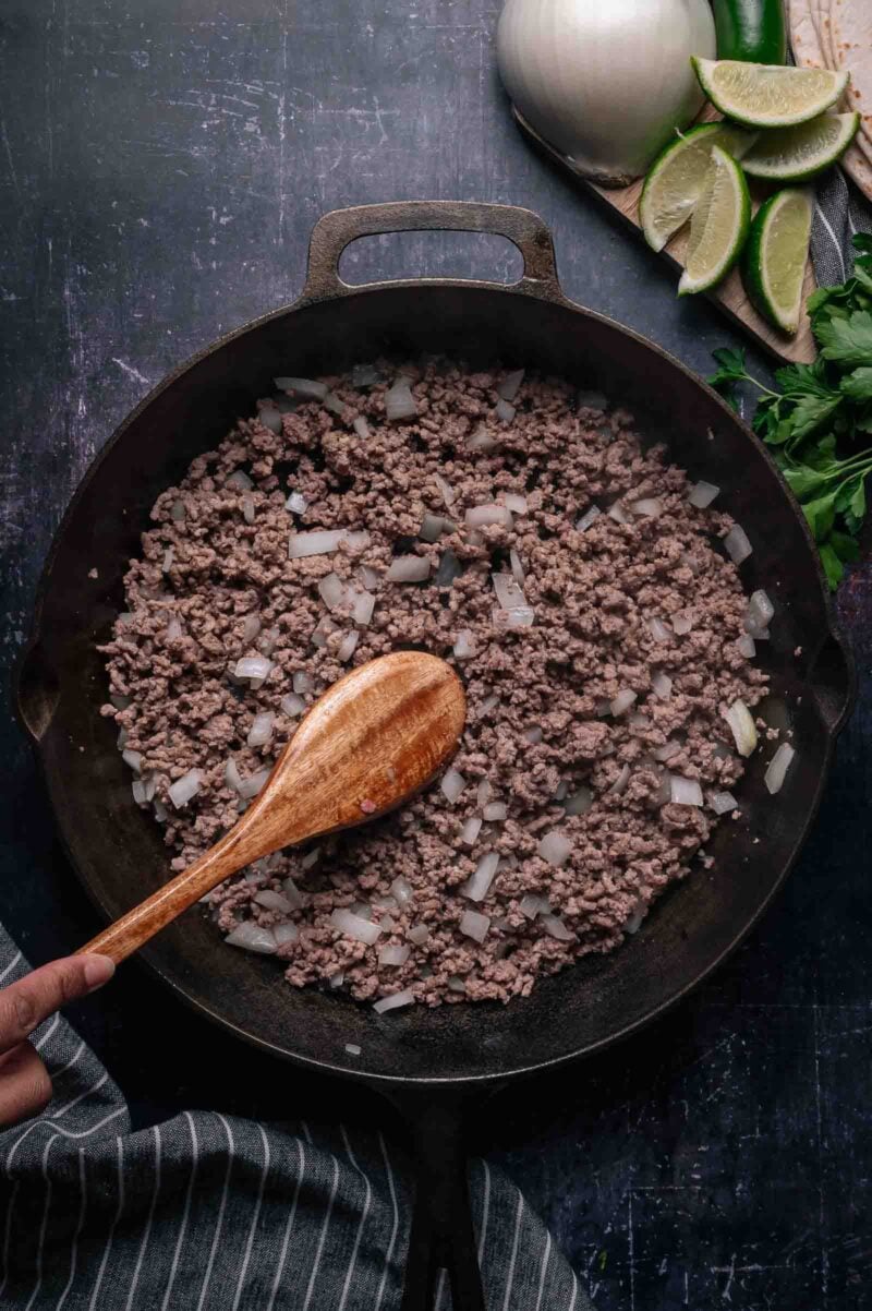 Ground beef and chopped onions being stirred in a cast iron skillet with a wooden spoon; limes, onion, and jalapeños on a cutting board nearby.