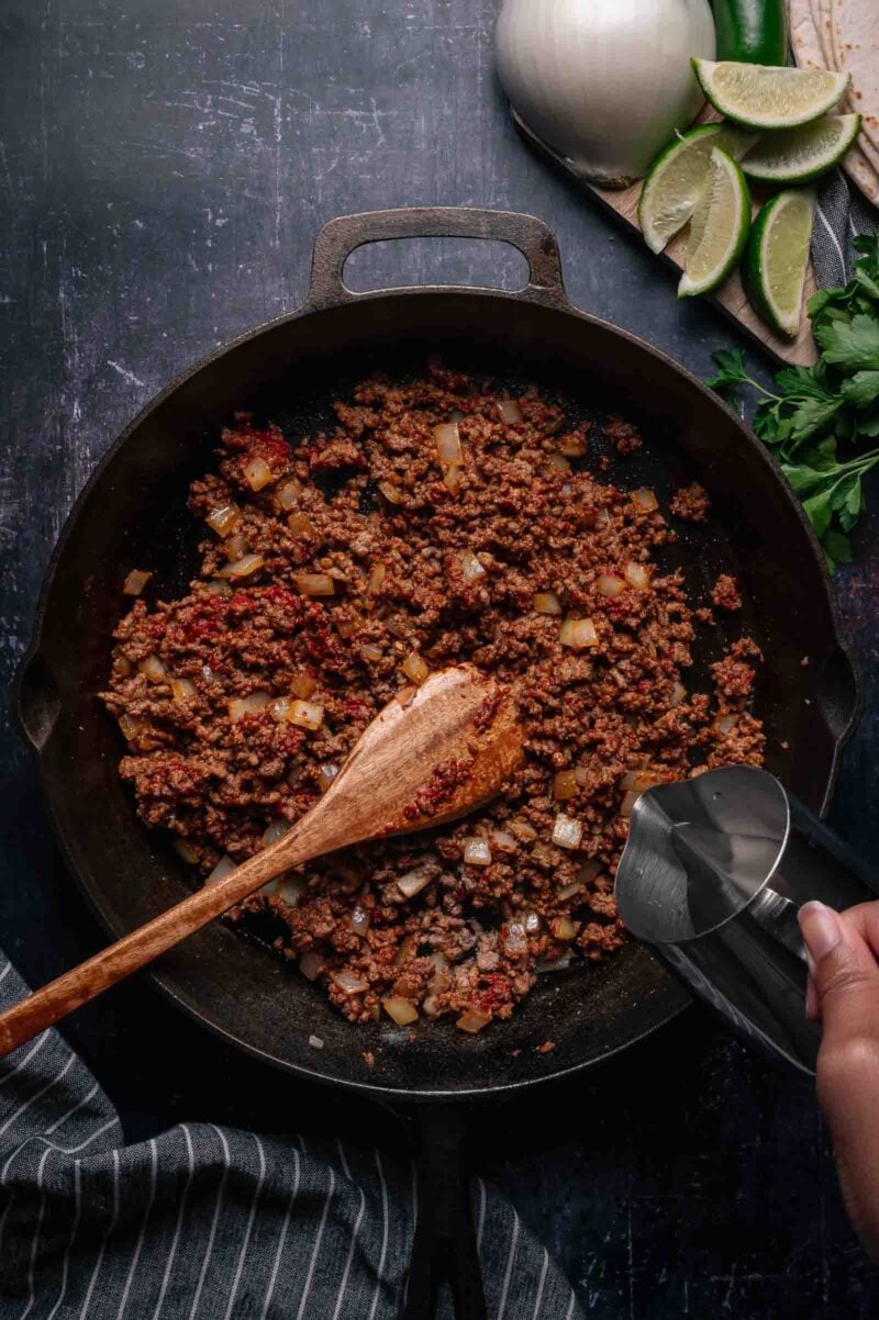 Ground beef and diced onions cooking in a cast iron skillet, with a hand pouring liquid from a metal cup; lime wedges, onion, and herbs are nearby.