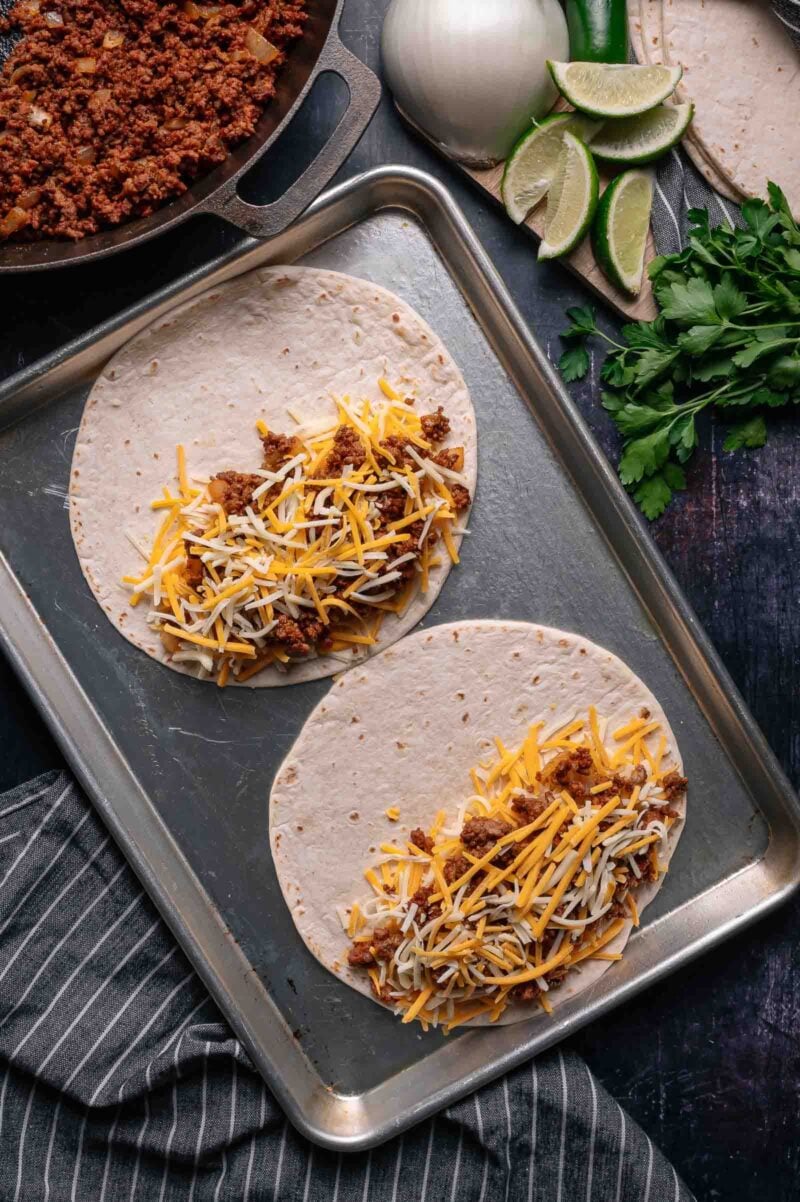 Two tortillas on a baking sheet, each topped with seasoned ground meat and shredded cheese, with a skillet, onion, lime wedges, jalapeño, and cilantro nearby.