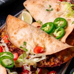 Three folded tacos filled with ground meat, shredded lettuce, diced tomatoes, and sliced jalapenos, served with lime wedges and extra jalapenos on the side.
