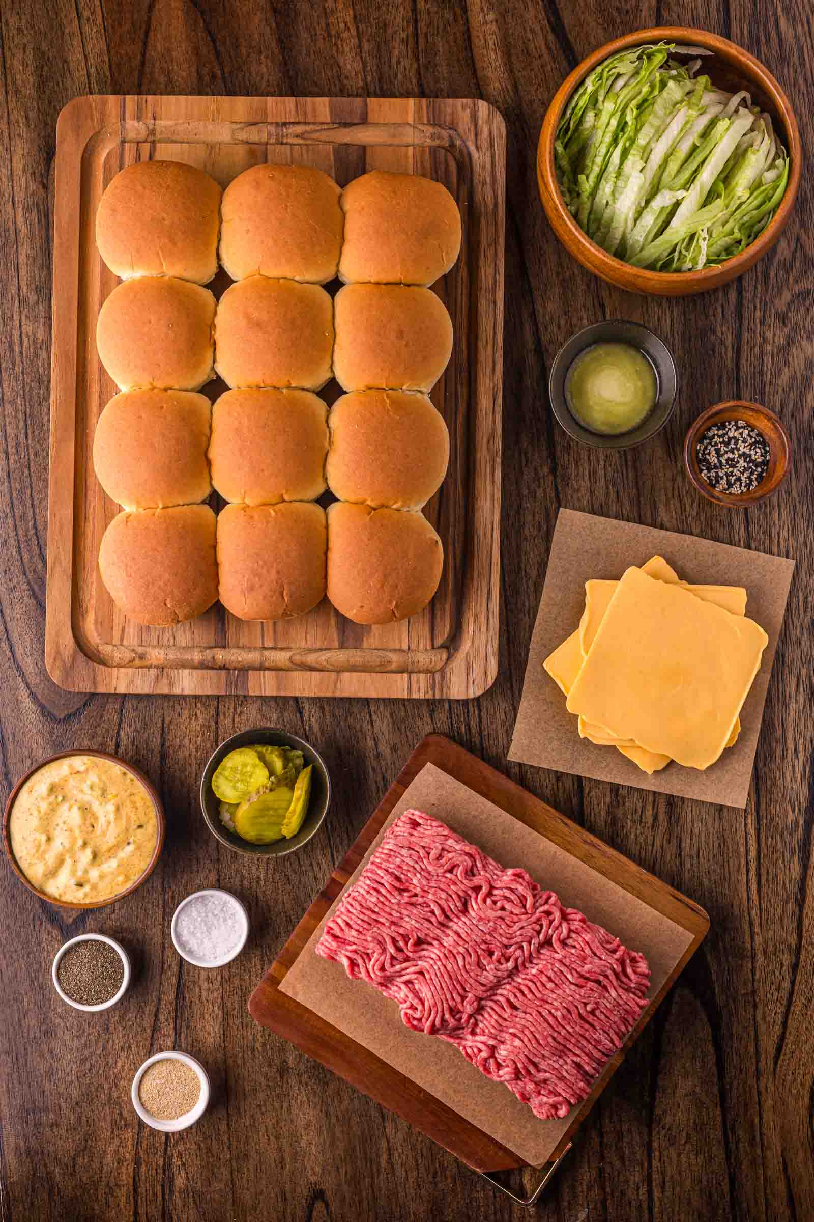 Top-down view of ingredients for big mac sliders, including slider buns, ground beef, cheddar cheese slices, shredded lettuce, pickles, sauces, and seasonings arranged on a wooden table.