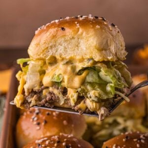 A spatula holds a Big Mac cheeseburger slider with lettuce, melted cheese, and sauce in a sesame seed-topped bun above a baking tray of similar sliders.