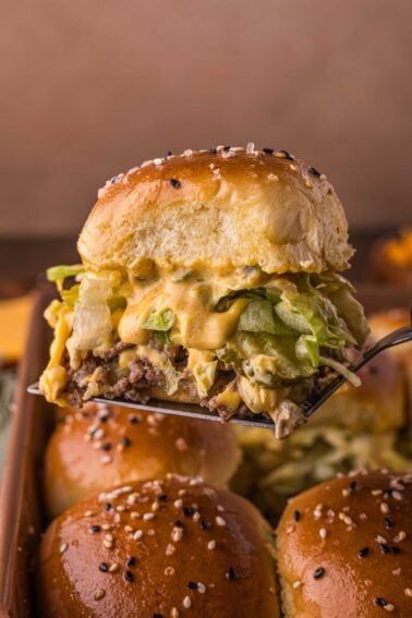 A spatula holds a Big Mac cheeseburger slider with lettuce, melted cheese, and sauce in a sesame seed-topped bun above a baking tray of similar sliders.